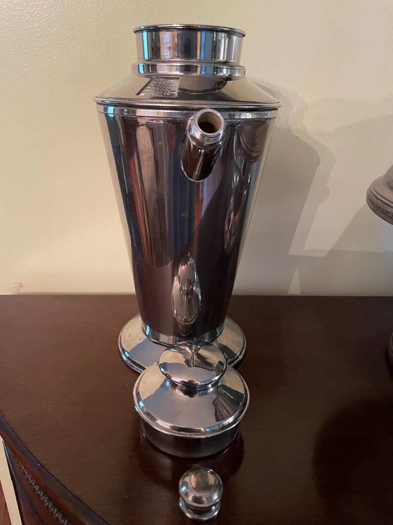 May include: A polished silver cocktail shaker with a conical body, a spout, and a lid. The shaker is displayed on a dark wooden surface. The lid and base are circular, and a small cap sits nearby. The design is vintage.