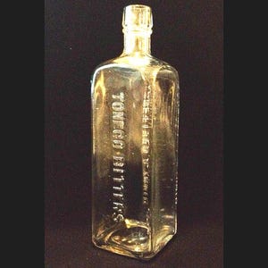May include: A clear glass bottle with a square shape and a glass stopper. The bottle is embossed with the text "Tonego Bitters".