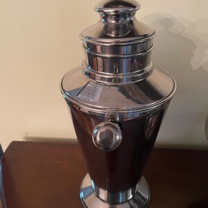 May include: A silver cocktail shaker with a dark brown body. The shaker has a rounded base, a bulbous midsection, and a lid with a small handle. A small, round knob is on the side. The shaker is on a dark brown surface.