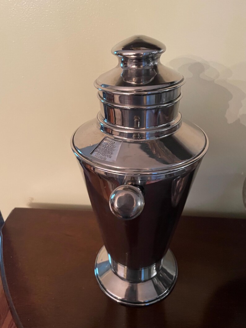 May include: A silver cocktail shaker with a dark brown body. The shaker has a rounded base, a bulbous midsection, and a lid with a small handle. A small, round knob is on the side. The shaker is on a dark brown surface.