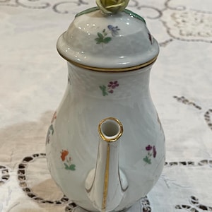 May include: A white porcelain teapot with a lid featuring a sculpted yellow rose finial. The body and lid are decorated with small, colorful hand-painted floral motifs and accented with thin gold trim along the spout and lid rim.