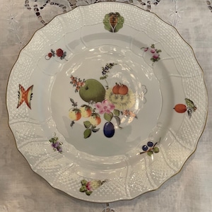May include: White porcelain plate with a scalloped edge and gold trim. The plate features a central floral and fruit design with a green apple, pink flowers, and various fruits. Additional floral and fruit motifs are scattered around the rim.