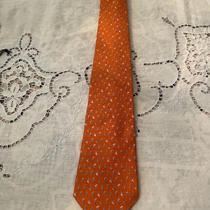 Peut inclure: Une cravate orange avec un motif r&eacute;p&eacute;t&eacute; de petites figures stylis&eacute;es blanches, bleues et jaunes. La cravate est faite d'un tissu lisse et est pr&eacute;sent&eacute;e sur une surface blanche &agrave; motifs. Un accessoire classique.
