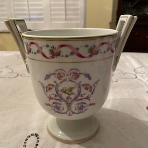 May include: White porcelain vase with two handles and a pedestal base. The vase is decorated with a floral and ribbon design in pink, yellow, and purple. Gold trim accents the rim, handles, and base.
