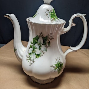 May include: White porcelain coffee pot with a floral design featuring white flowers and green leaves. The pot has a gold trim around the lid and base.