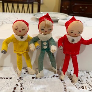 May include: Three vintage elf dolls with painted faces, wearing red, yellow, and green felt outfits. Each doll has a red hat, white collar, and silver glitter accents on the cuffs. The dolls are standing on a white lace tablecloth.