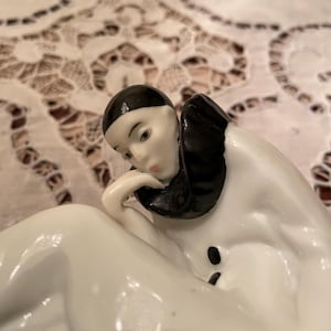 May include: A porcelain figurine of a reclining figure in a white outfit with a black collar and black hair. The figure is resting their chin on their hand. The figurine has a smooth, glossy finish and is set against a patterned background.