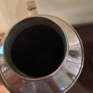 May include: A silver metal pitcher with a wide, circular opening and a black interior. The pitcher has a handle and a spout, and it appears to be made of a shiny, reflective material. The pitcher is likely used for serving beverages.