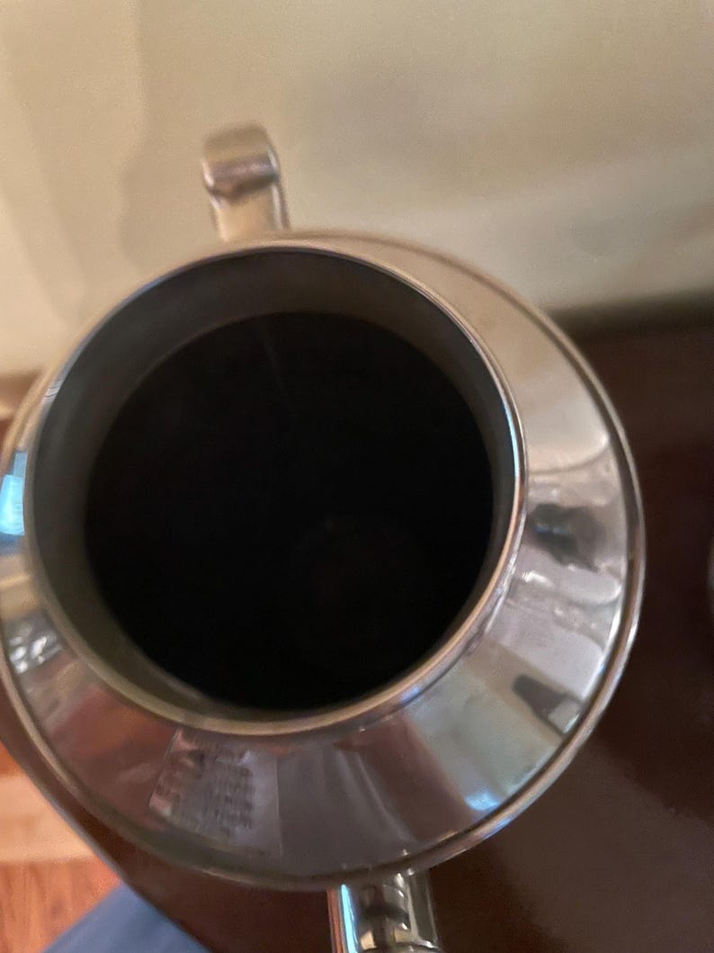 May include: A silver metal pitcher with a wide, circular opening and a black interior. The pitcher has a handle and a spout, and it appears to be made of a shiny, reflective material. The pitcher is likely used for serving beverages.