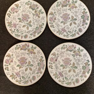 May include: A set of four round white ceramic plates featuring a decorative floral pattern in shades of pink, yellow, and green with intricate foliage details. Each plate has a thin gold-toned rim around the edge, creating a uniform appearance.