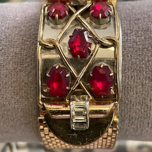 May include: A vintage gold-tone bracelet with a rectangular centerpiece. The centerpiece features five large, faceted red gemstones and a decorative gold filigree design. The bracelet has a mesh-like band.