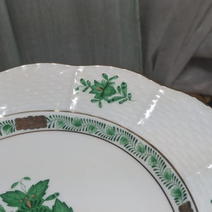 May include: A white porcelain plate with a green and gold floral design. The plate has a scalloped edge and a raised rim.