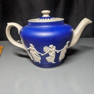 May include: A blue and white teapot with a white relief design of figures dancing around a central figure. The teapot has a white lid with a decorative border.
