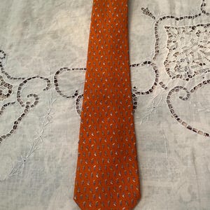 Peut inclure: Une cravate orange avec un motif r&eacute;p&eacute;t&eacute; de petites figures stylis&eacute;es bleues, blanches et jaunes. La cravate est faite d'un tissu lisse et est pr&eacute;sent&eacute;e sur une surface blanche &agrave; motifs.