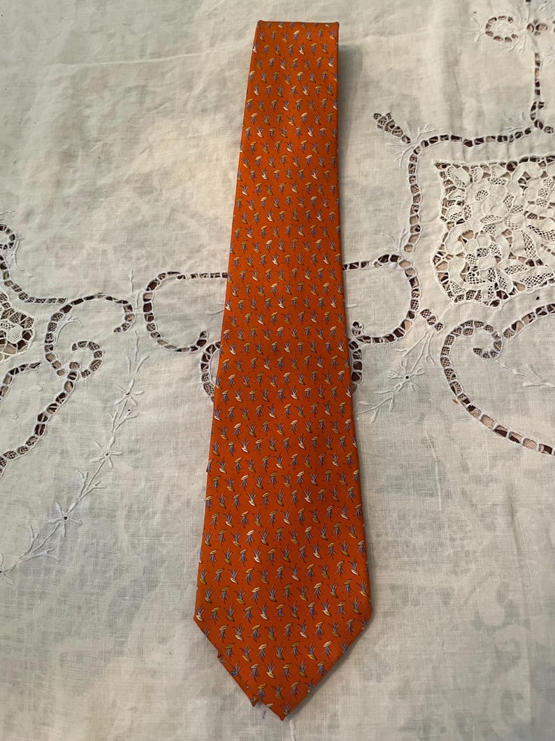 Peut inclure: Une cravate orange avec un motif r&eacute;p&eacute;t&eacute; de petites figures stylis&eacute;es bleues, blanches et jaunes. La cravate est faite d'un tissu lisse et est pr&eacute;sent&eacute;e sur une surface blanche &agrave; motifs.