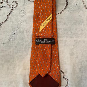 Peut inclure: Une cravate en soie orange avec un motif r&eacute;p&eacute;t&eacute; de petites formes de feuilles stylis&eacute;es en bleu, blanc et jaune. La cravate a une doublure marron fonc&eacute; et une &eacute;tiquette indiquant "Salvatore Ferragamo, Made in Italy."