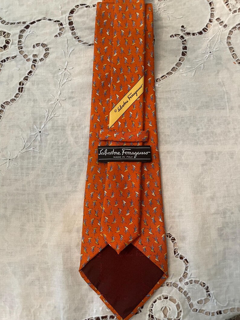 Peut inclure: Une cravate en soie orange avec un motif r&eacute;p&eacute;t&eacute; de petites formes de feuilles stylis&eacute;es en bleu, blanc et jaune. La cravate a une doublure marron fonc&eacute; et une &eacute;tiquette indiquant "Salvatore Ferragamo, Made in Italy."