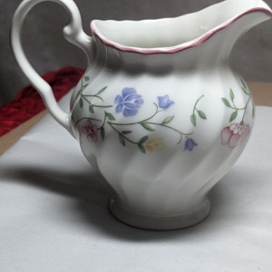 May include: White ceramic creamer with a floral design. The creamer has a curved handle and a spout. The rim is trimmed with a pink border. The floral pattern includes blue, pink, and yellow flowers with green stems and leaves.