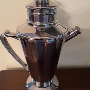 May include: A polished silver cocktail shaker with a handle and a spout. The shaker has a tiered lid and a wide base. The handle curves gracefully from the body. The overall design is sleek and vintage.
