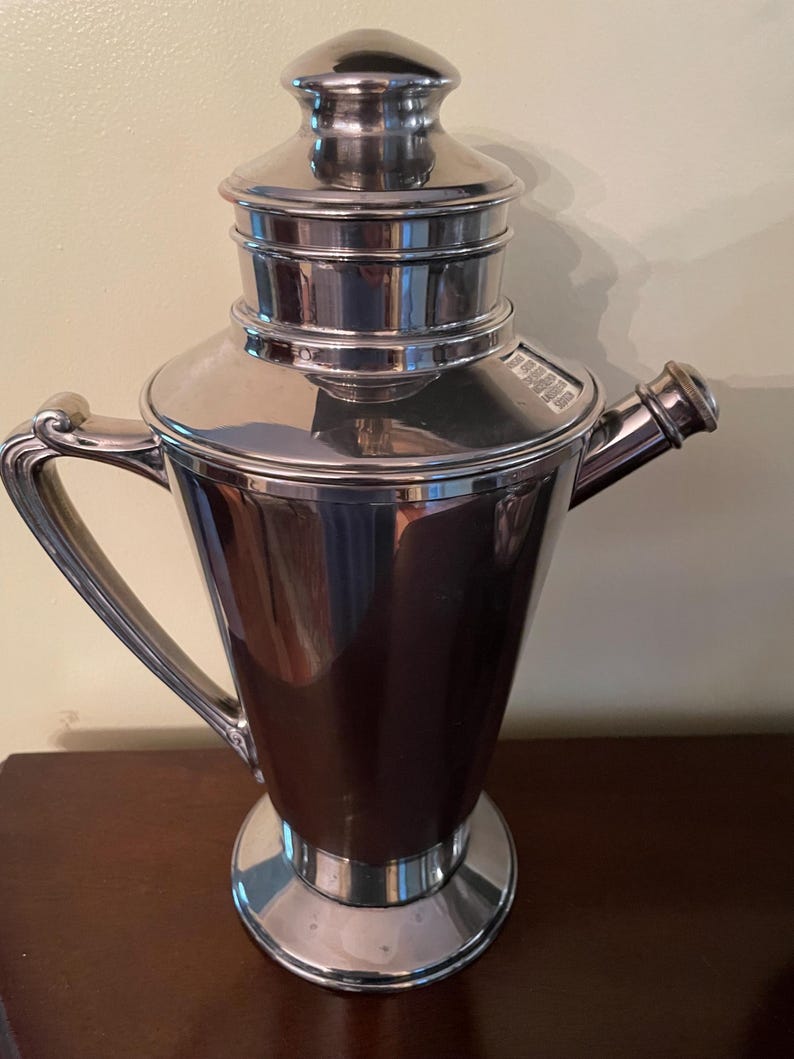 May include: A polished silver cocktail shaker with a handle and a spout. The shaker has a tiered lid and a wide base. The handle curves gracefully from the body. The overall design is sleek and vintage.