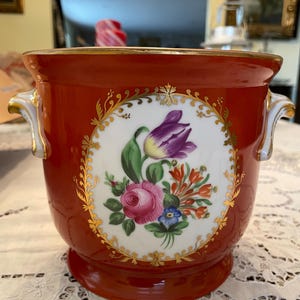 May include: A red ceramic planter with a white circular design featuring a floral arrangement of pink roses, purple tulips, and other colorful flowers. The planter has gold trim and handles.