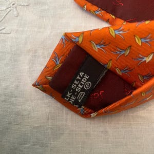 Peut inclure: Cravate en soie orange avec un motif floral r&eacute;p&eacute;t&eacute; bleu et jaune. La cravate a une doublure bordeaux fonc&eacute; et une &eacute;tiquette qui indique "LK-SETA DIE-SEIDE".