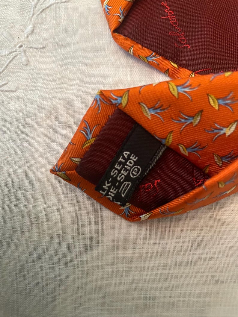 Peut inclure: Cravate en soie orange avec un motif floral r&eacute;p&eacute;t&eacute; bleu et jaune. La cravate a une doublure bordeaux fonc&eacute; et une &eacute;tiquette qui indique "LK-SETA DIE-SEIDE".