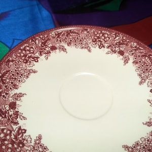 May include: A white ceramic saucer with a red floral pattern around the edge. The saucer has a small, white center depression.