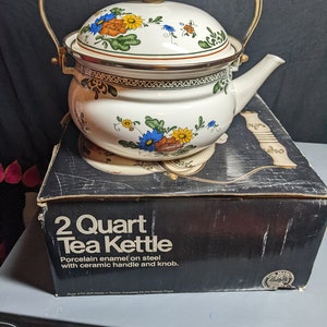 May include: A white 2 quart tea kettle with a floral design and a ceramic handle and knob. The kettle is sitting on a black box with the words "2 Quart Tea Kettle" printed on it.