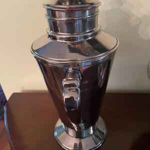May include: A polished silver cocktail shaker with a handle and a lid. The shaker has a conical body and a rounded top. The handle is rectangular and attached to the side. The lid is silver and has a small knob on top. The shaker is made of metal.
