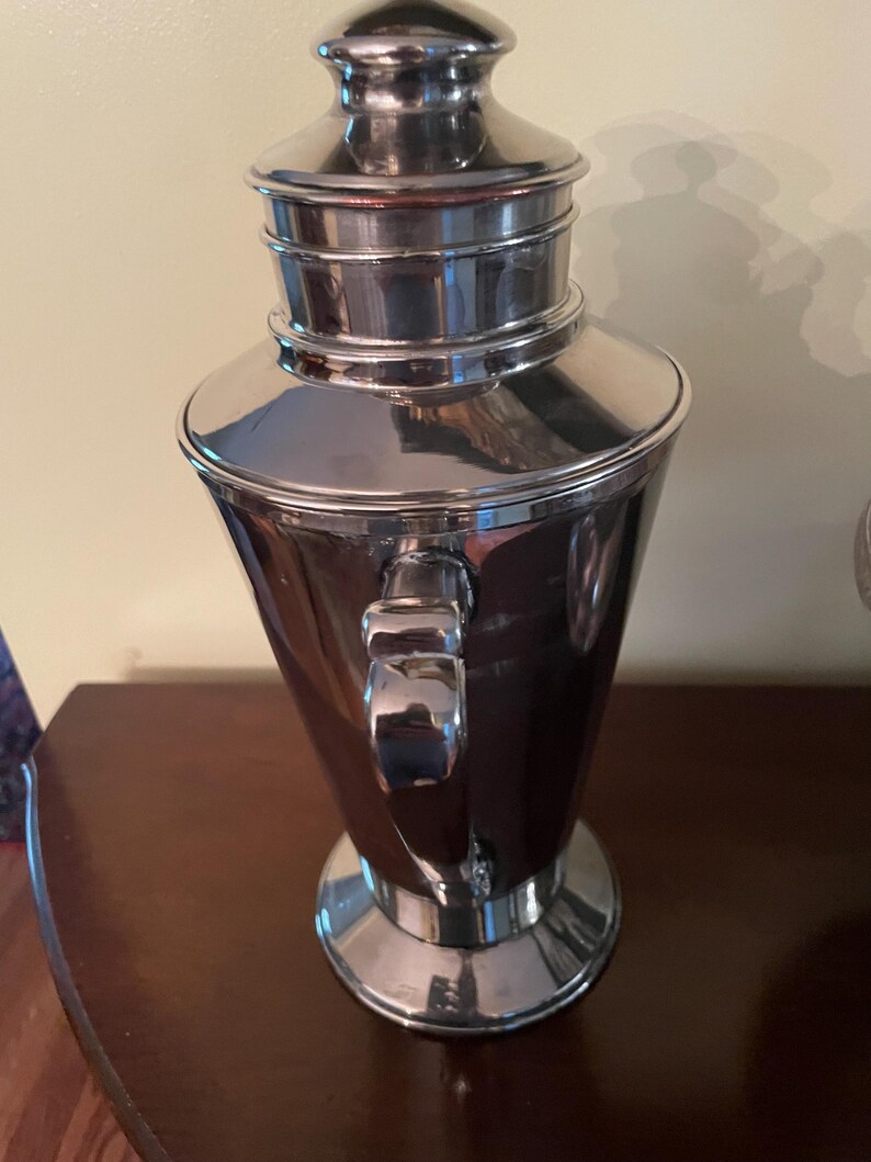 May include: A polished silver cocktail shaker with a handle and a lid. The shaker has a conical body and a rounded top. The handle is rectangular and attached to the side. The lid is silver and has a small knob on top. The shaker is made of metal.