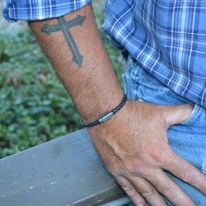 May include: A black braided leather bracelet with a silver magnetic clasp. The bracelet is worn on a wrist with a cross tattoo and is paired with a blue plaid shirt and blue jeans.