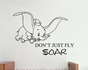 Puede incluir: Pegatina de pared en blanco y negro con Dumbo el elefante volando con el texto "Don't just fly SOAR". La pegatina se muestra en una guardería con una cuna blanca y un sillón azul.