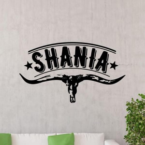 May include: Black and white vinyl wall decal with the name "Shania" in a western-style font with a star on each side of the name. The name is above a stylized black bull's head with horns.