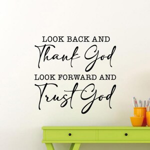 May include: A wall decal with the text "LOOK BACK AND Thank God LOOK FORWARD AND Trust God" in black script. The decal is on a white wall above a green table with orange containers holding pencils and pens.