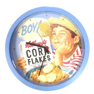 Op de afbeelding: Blauwe ronde klok met een afbeelding van een jongen die Kellogg's Corn Flakes eet. De wijzerplaat van de klok heeft een witte achtergrond met zwarte cijfers en wijzers. De jongen draagt een strohoed en een rood-wit gestreept shirt. De tekst "Boy!" staat in een spreekballon boven het hoofd van de jongen.