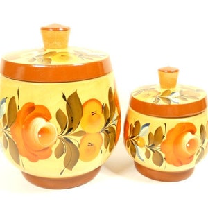 Op de afbeelding: Twee houten voorraadpotten met deksels, beschilderd met een bloemmotief in tinten oranje, geel en groen. De potten zijn gemaakt van hout en hebben een traditionele Russische volkskunststijl.
