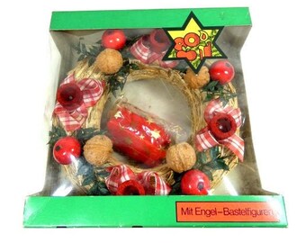 May include: A straw wreath with red and white ribbon, red apples, and nuts. The wreath is in a clear plastic box with a green background. The box has the text "Mit Engel-Bastelfiguren" on the bottom.