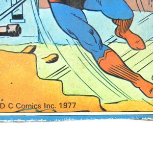 May include: A comic book cover featuring a superhero in a blue and red suit flying through the air. The cover is yellow and green with the text "DC Comics Inc. 1977".
