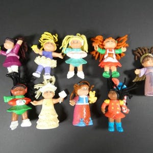 May include: A collection of ten vintage Cabbage Patch Kids figurines. The dolls are dressed in various outfits, including dresses, skirts, and angel costumes. The dolls have different hair colours and styles, and some are holding accessories.