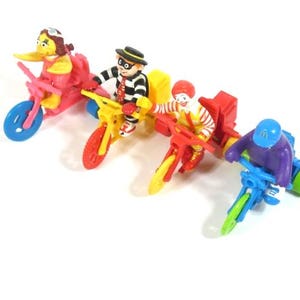 Puede incluir: Un conjunto de cuatro figuras de juguete de plástico de colores que montan triciclos. Las figuras incluyen un pájaro amarillo, una figura a rayas con un sombrero negro, una figura a rayas rojas y blancas y una figura morada con un casco azul.