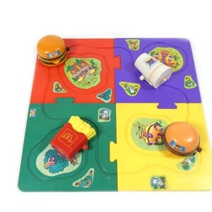 Può includere: Un colorato puzzle per bambini con quattro sezioni ad incastro in rosso, verde, viola e giallo. Il puzzle include versioni giocattolo di un hamburger, patatine fritte, una bevanda e una campana. Il puzzle ha un tema McDonald's.