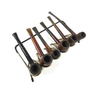 Op de afbeelding: Een set van zeven houten pijpen met zwarte kommen, tentoongesteld op een metalen rek. De pijpen zijn in verschillende bruintinten en hebben een rustieke uitstraling.