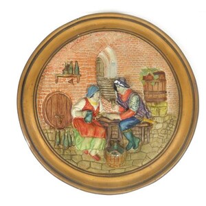Può includere: Una targa da parete in ceramica con una scena 3D di due persone sedute a un tavolo in un edificio di mattoni. La scena è dipinta in colori caldi con dettagli come una botte, bottiglie e un cesto di frutta.