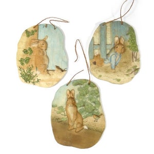 May include: Three decorative wooden plaques with rabbit illustrations. Each plaque features a different scene with a rabbit, painted in soft pastel colors. The plaques have a rustic, aged appearance and are strung with twine for hanging.