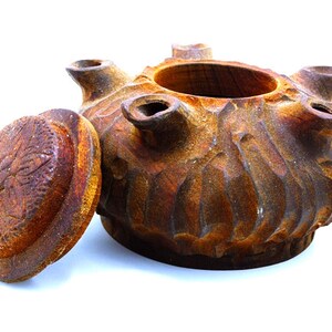 May include: A rustic brown ceramic pot with a lid, featuring a textured, wavy design. The pot has five openings around the top, and the lid has a raised, circular design.