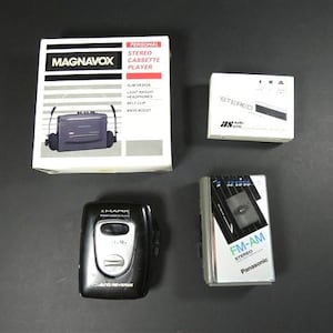 Könnte beinhalten: Eine Auswahl an Vintage-Stereo-Kassettenplayern und Radios. Enthält einen Magnavox-Player in der Originalverpackung, ein kleines weißes Stereo, einen schwarzen Smark-Player und ein silbernes Panasonic FM-AM-Stereo-Radio.