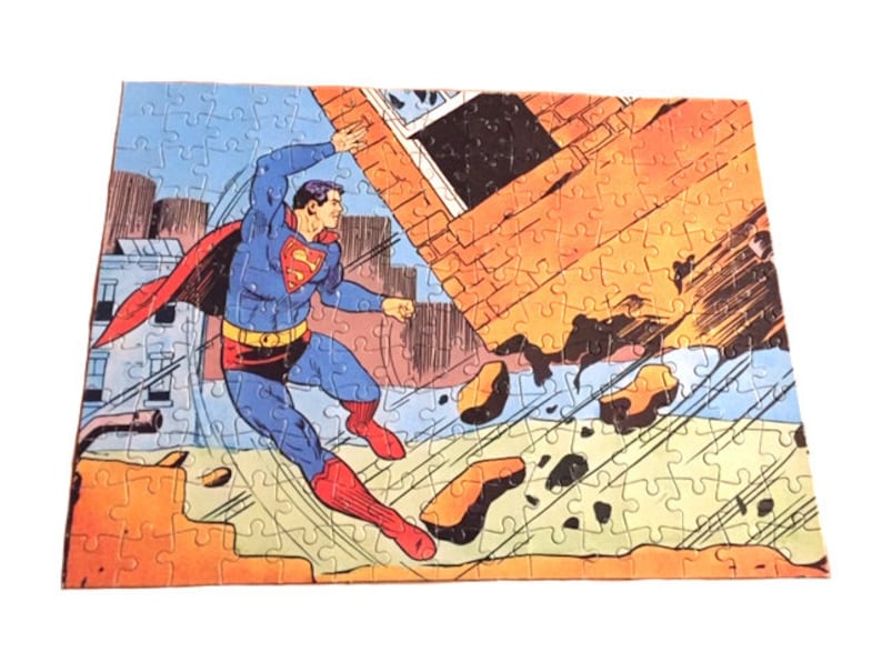 May include: A vintage jigsaw puzzle featuring a comic book illustration of Superman flying through the air, breaking through a brick wall. The puzzle is made of cardboard and has a colorful design.