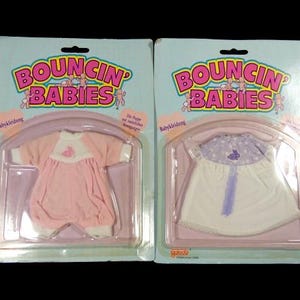 May include: Two packages of doll clothes for a baby doll. The packages are pink and blue and feature the text "BOUNCIN' BABIES". One package has a pink dress with a white trim and a bunny design. The other package has a white dress with a purple trim and a bunny design.