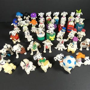 Könnte beinhalten: Eine Sammlung von Dalmatiner-Figuren in verschiedenen Posen. Die Figuren sind weiß mit schwarzen Flecken, einige tragen farbige Accessoires. Ein Fußball ist ebenfalls vorhanden. Dunkler Hintergrund.